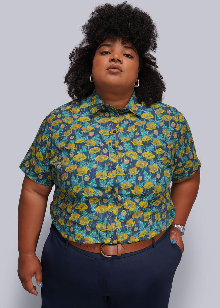 QX=A plus-size model wearing a short-sleeve button-up shirt with a yellow and teal floral pattern, paired with navy blue pants and a brown belt, against a solid gray background.