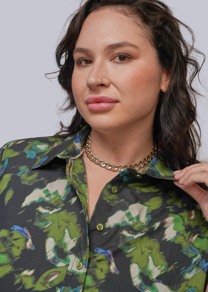 1X=A close-up of a woman wearing a dark button-up shirt with a green, beige, and blue abstract print, styled with a chunky gold chain necklace against a light gray background.