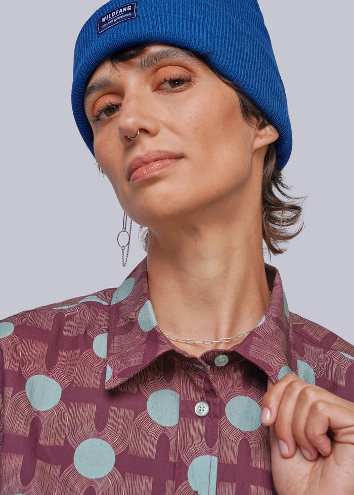 S=A close-up of a person wearing a bright blue ribbed knit beanie with a small black brand patch that reads 'WILDFANG' and a patterned button-up shirt in mauve and light blue.