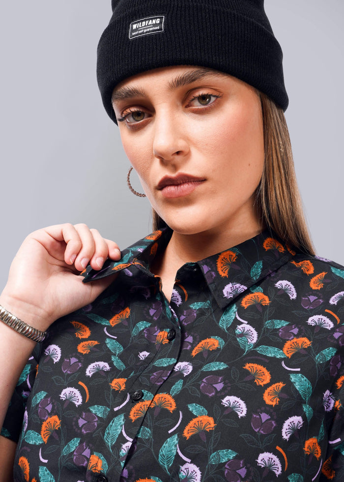 S= A person in a black beanie and hoop earrings looks at the camera with a neutral expression. They wear The Essential Button Up by Wildfang, featuring a vibrant floral pattern in purple, orange, and green on black, offering breathable cotton comfort.