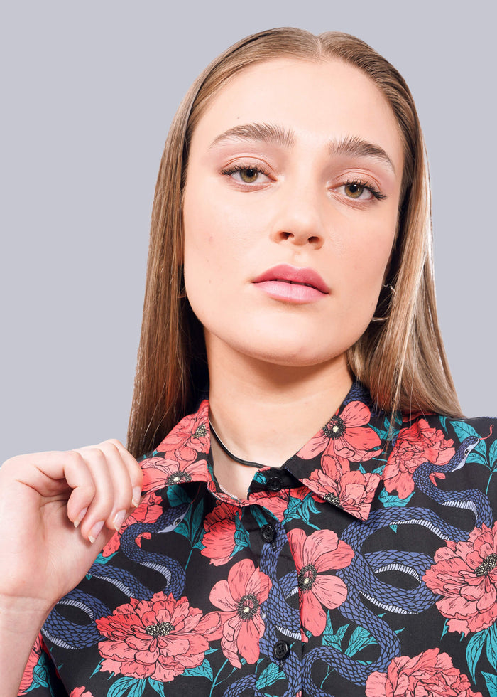 S=A young woman with straight, light brown hair wears The Essential Button Up, featuring pink floral and blue snake patterns. She holds the collar with one hand and looks confidently at the camera against a white background.