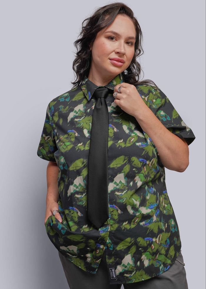 1X=A woman modeling a short-sleeve button-up shirt with a green tropical bird print, styled with a black necktie and gray trousers against a solid gray studio background.