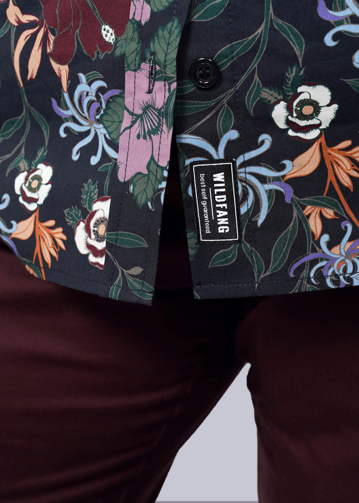 1X=Close-up of a person wearing maroon pants and The Essential Button Up, featuring a vibrant Wildfang print. A WILDFANG brand tag is visible by the hem, showcasing the shirt’s tailored fit and attention to detail.