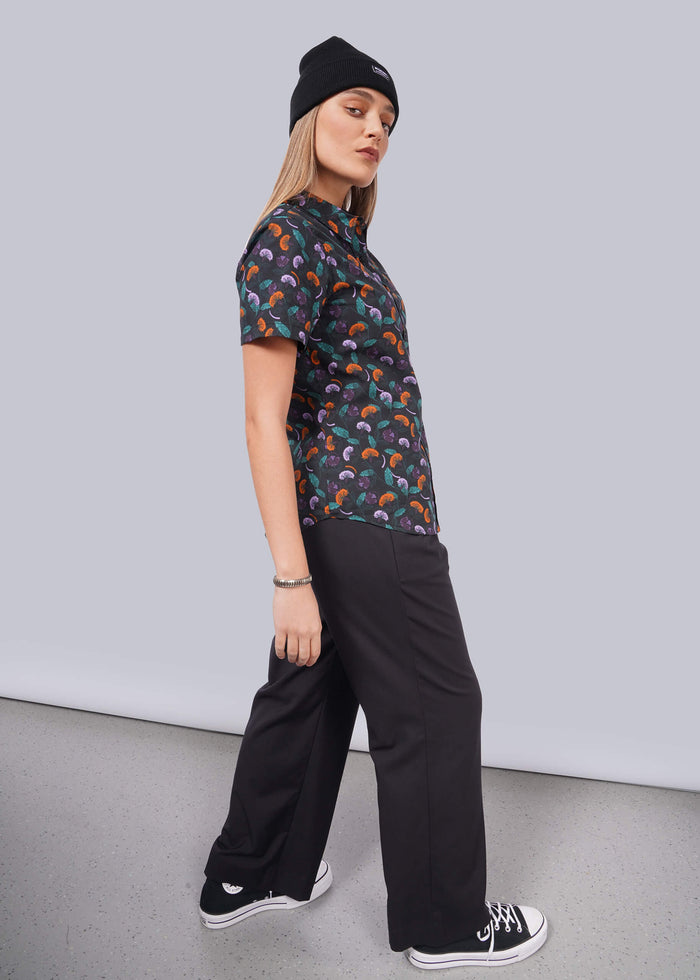 S=A person confidently wears a black Essential Button Up by Wildfang in breathable cotton with a colorful pattern, complemented by black wide-leg pants, shoes, and beanie. Theyre looking to the side against a plain white backdrop.
