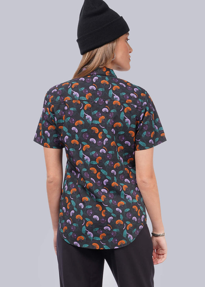 S=A person wearing a black beanie, a short-sleeved Essential Button Up with a colorful abstract pattern, tailored fit black pants, and sneakers stands facing away from the camera against a plain light background.