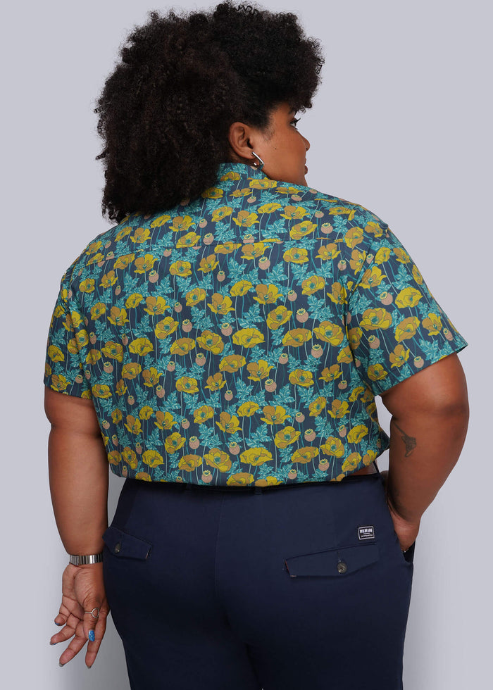 1X=A person shown from the back wearing a short-sleeve button-up shirt with a blue, yellow, and green floral pattern, paired with navy pants, against a light gray studio background.