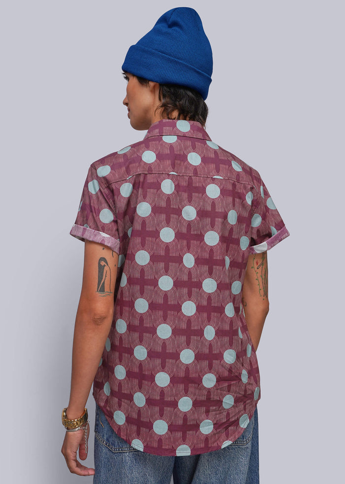 S=A person shown from the back wearing a short-sleeve button-up shirt with a burgundy base and a white geometric cross-and-dot pattern, paired with blue jeans and a blue beanie, against a solid gray studio background.