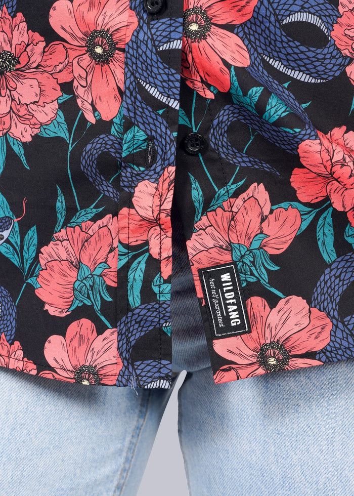 S=A close-up of The Essential Button Up by Wildfang, featuring pink flowers and blue snakes on black cotton. Partially unbuttoned and styled with light blue jeans, its tailored fit offers stylish comfort. Label reads WILDFANG.