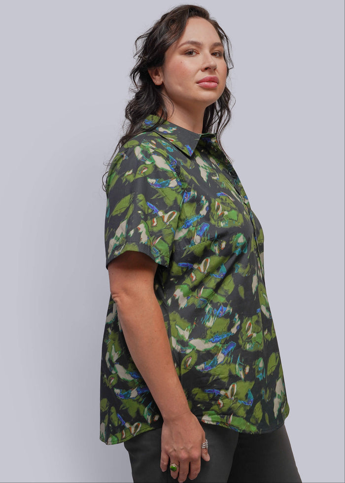 1X=A woman wearing a short-sleeve button-up shirt with a green, blue, and white tropical leaf pattern on a dark background, photographed against a solid light gray studio backdrop.