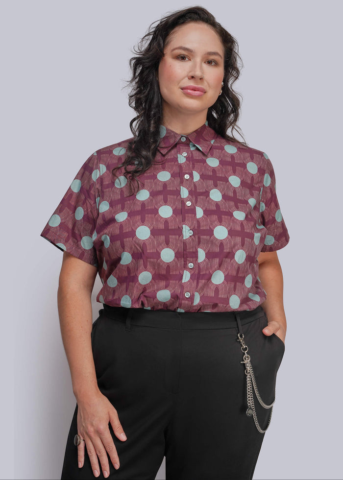1X=A woman modeling a short-sleeve button-up shirt with a purple and light blue geometric pattern, paired with black trousers, against a solid gray studio background.