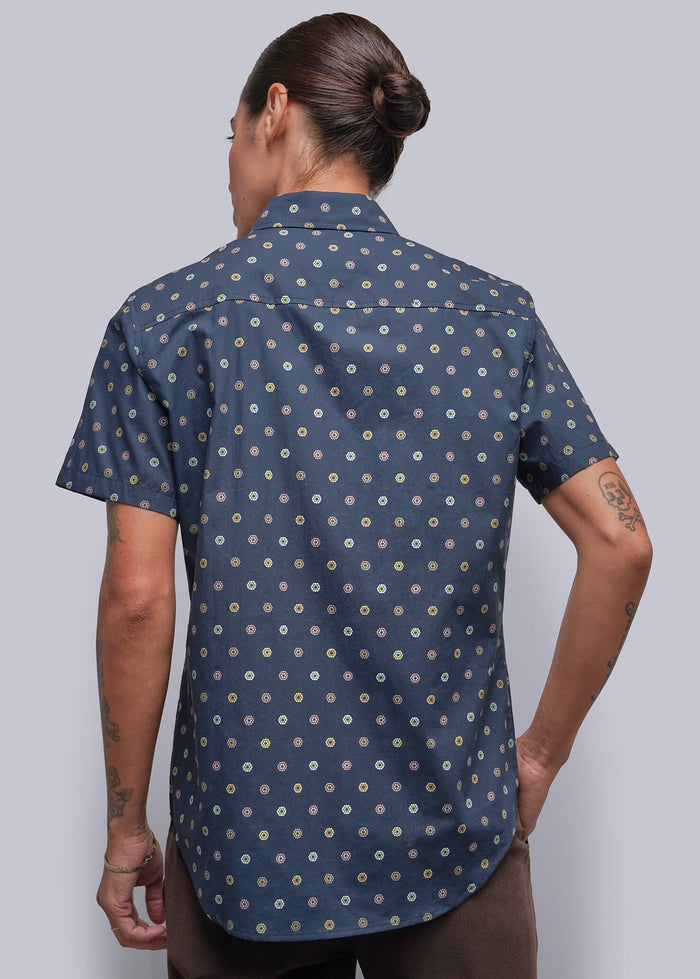 S=A person viewed from the back wearing a navy blue short-sleeve button-up shirt with a small white and yellow floral pattern, paired with brown trousers, against a solid gray background.