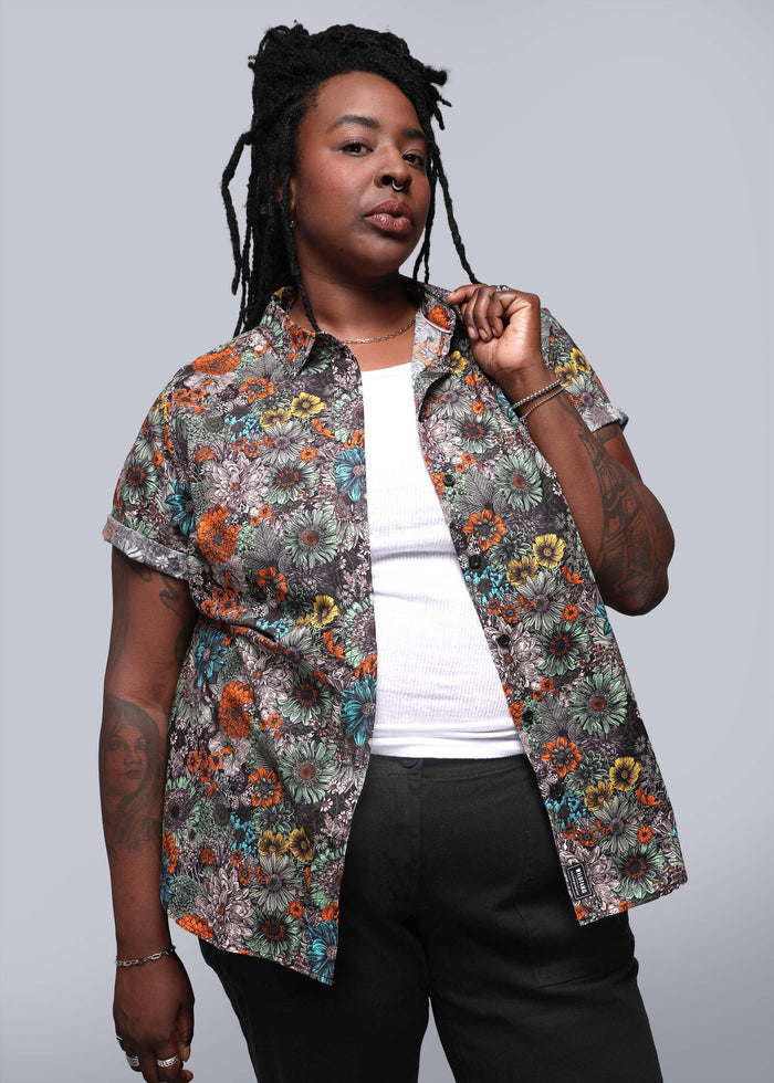 1X=Model wears white shirt under floral printed short sleeved button up shirt and black pants in front of grey background.