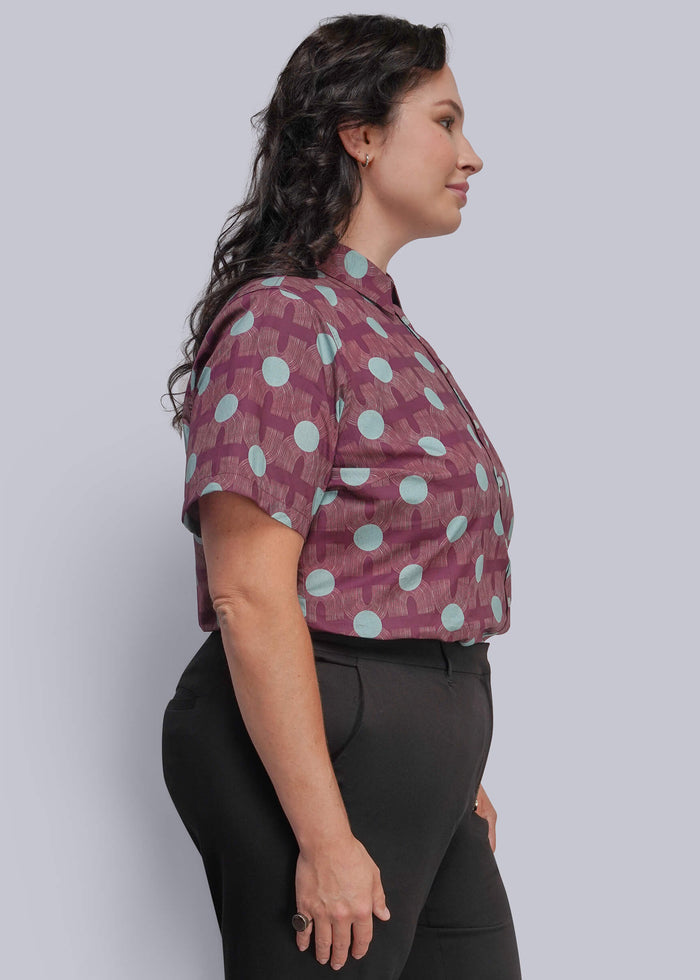1X=A woman in side profile wearing a short-sleeve button-up shirt with a purple and light blue geometric pattern, paired with black pants, against a solid light gray background.