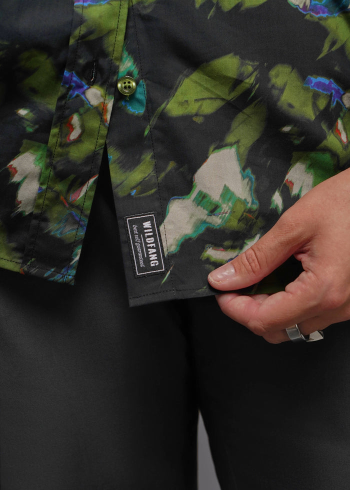 1X=Close-up of a person wearing a button-front shirt with a dark abstract print in green, blue, white, and black, paired with black pants. A hand is pulling the shirt hem to reveal a black brand label that reads WILDFANG.