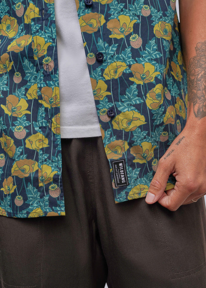 S=Close-up of a person wearing a short-sleeve button-front shirt with a dark teal background and a yellow floral print, layered over a white t-shirt and paired with dark brown pants. A black brand label reading 'SILKSTITCH' is visible near the hem.