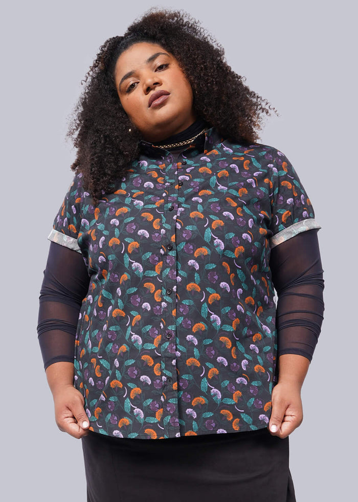 1X= A person with curly hair stands confidently against a white background, wearing The Essential Button Up featuring Wildfang branded buttons. Over a dark long-sleeve top, a black skirt, and black boots complete the tailored fit ensemble. The pose is relaxed yet self-assured.