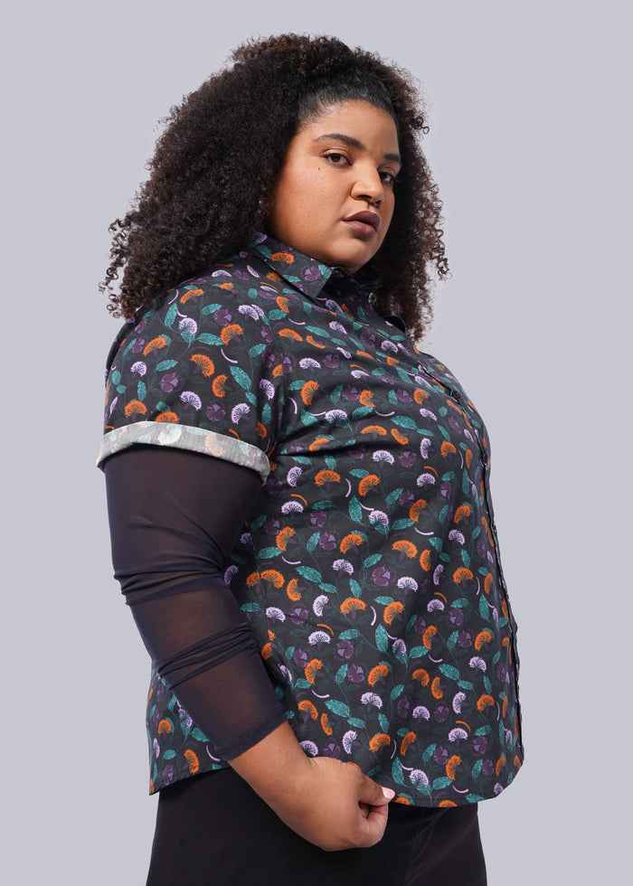 1X= A person with curly hair confidently poses against a plain background, wearing The Essential Button Up from Wildfang. This colorful, patterned shirt, made from breathable cotton for comfort, features branded buttons, rolled-up sleeves, and a tailored fit revealing a sheer black top underneath.