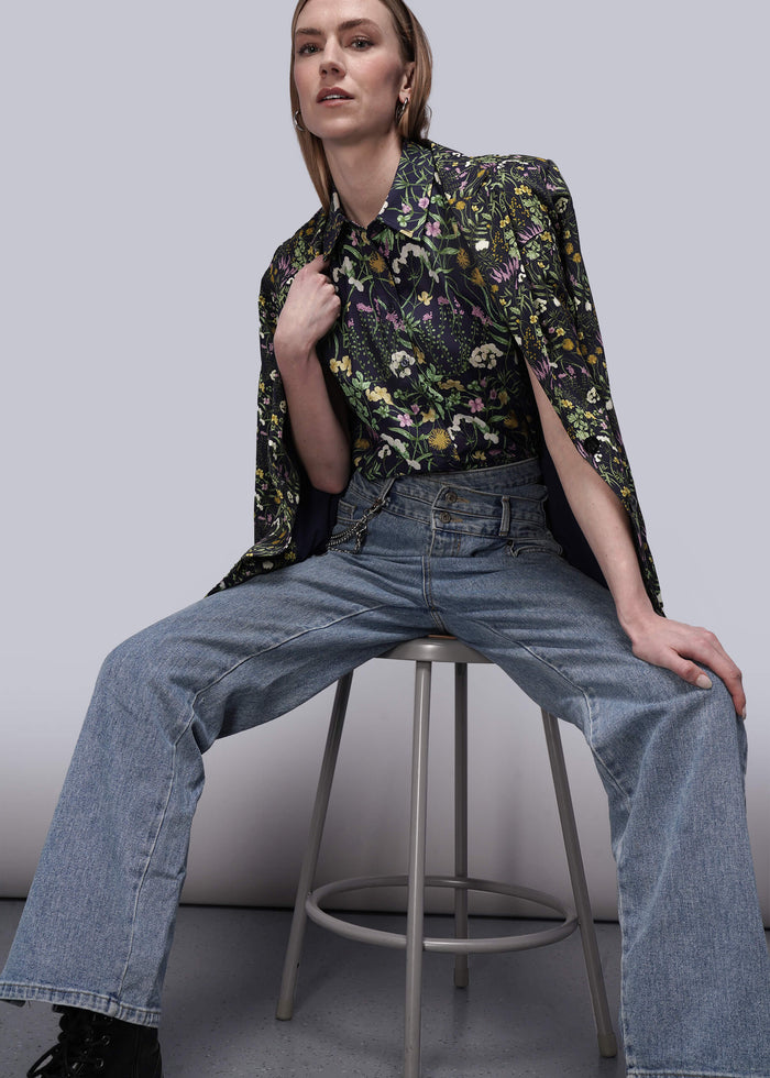 S=Person wearing a floral shirt, blazer, and jeans sitting on a stool against a plain background