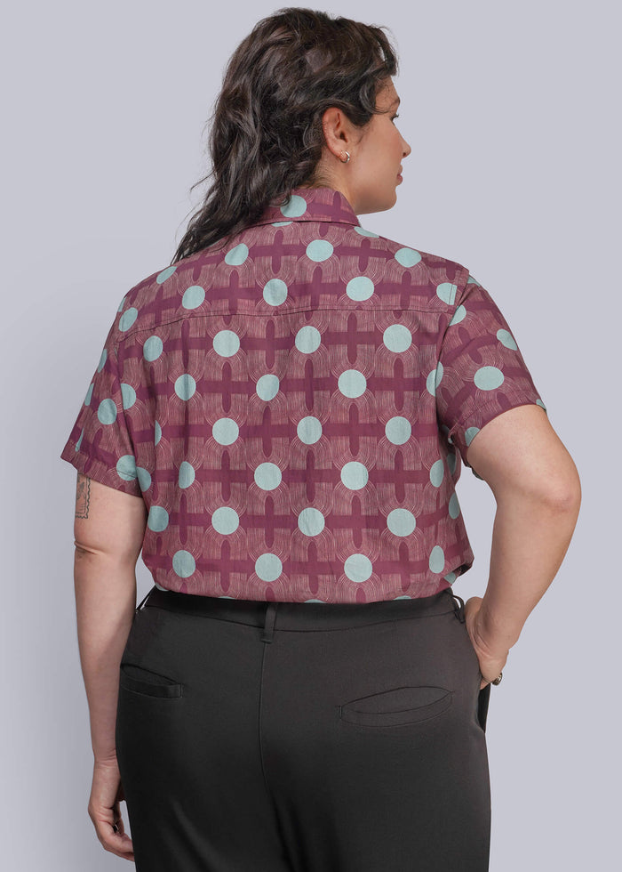 1X=A woman shown from the back wearing a short-sleeve button-up shirt with a burgundy and light blue geometric pattern, paired with black trousers, against a solid gray studio background.