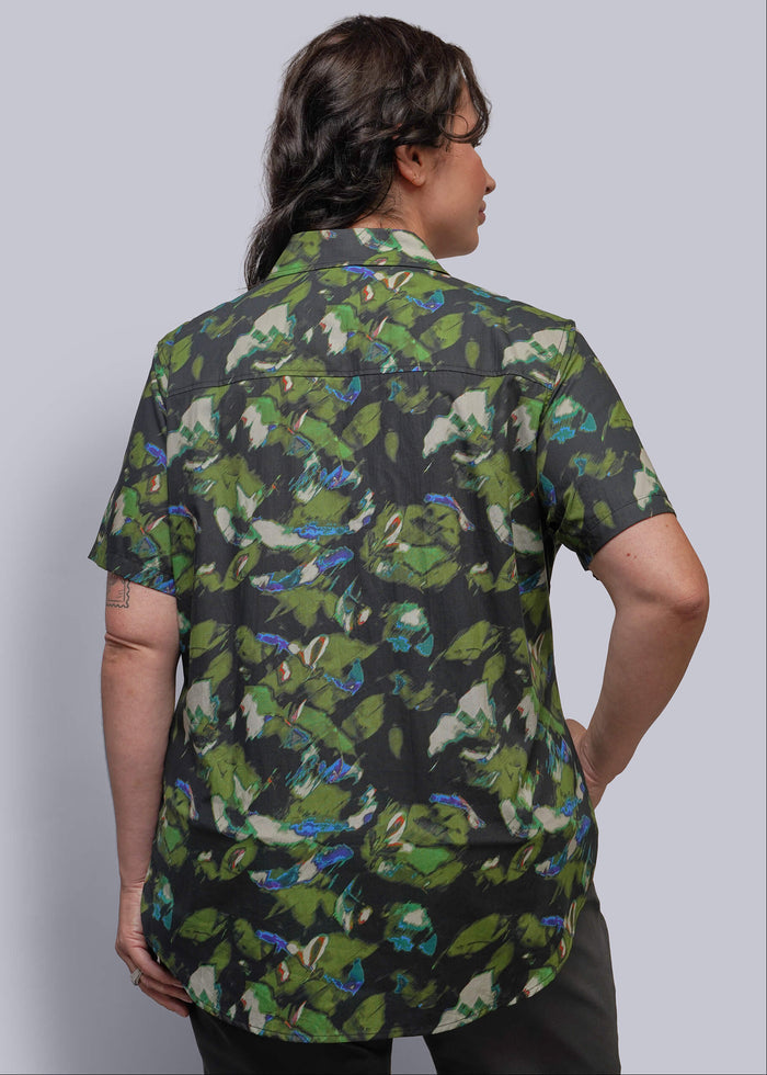 1X=A person shown from the back wearing a short-sleeve button-up shirt with an abstract camouflage-style print in green, black, blue, and white, against a plain light gray studio background.