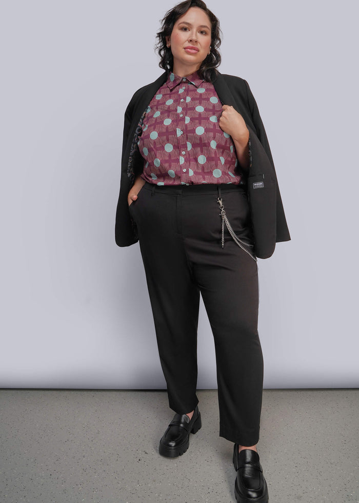 1X=A woman modeling a black blazer, black trousers, and a patterned button-up shirt with a burgundy and light blue polka dot design, standing against a light gray studio background.