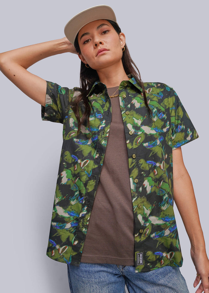 S=A model wearing a green tropical parrot print short-sleeve button-up shirt over a brown t-shirt, blue jeans, and a beige cap, set against a solid gray studio background.