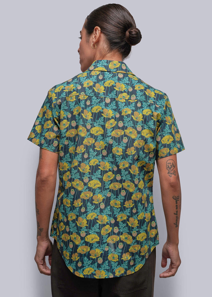 S=A person viewed from the back wearing a short-sleeve button-up shirt with a dense yellow and teal floral pattern on a dark background, paired with dark trousers, set against a solid gray studio backdrop.