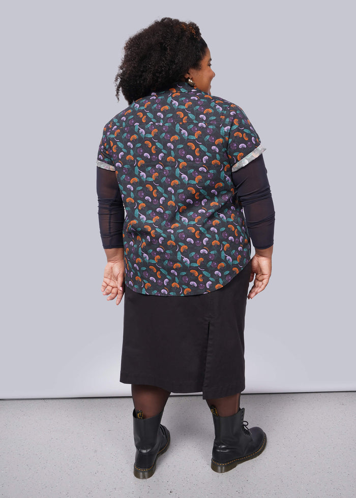 1X=A person with curly hair stands against a white wall, wearing The Essential Button Up, a colorful patterned, breathable cotton shirt. They pair it with a black skirt and boots, arms relaxed at their sides. The background is simple white with gray flooring.