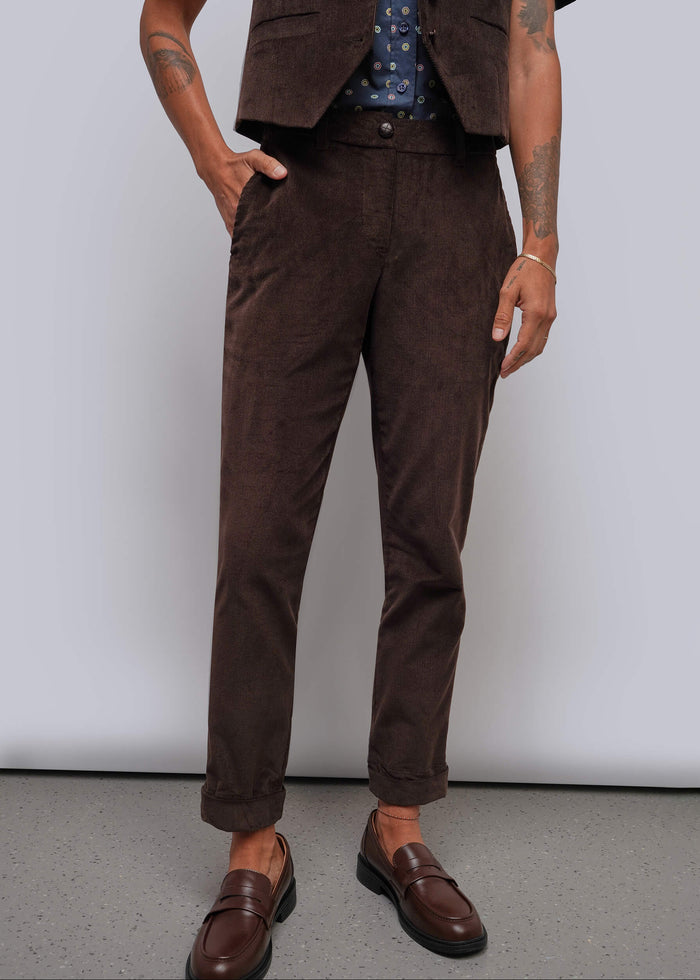 S=A person wearing dark brown corduroy trousers, styled with a matching brown vest and brown leather loafers against a plain light gray studio background.