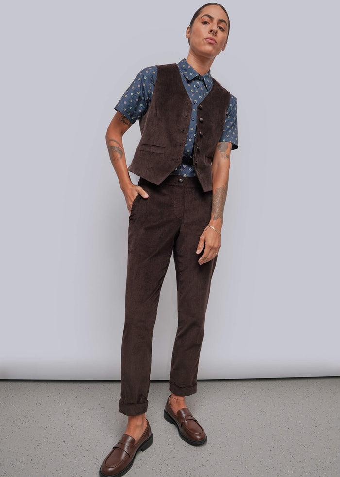 S=A model wearing a dark brown corduroy vest and matching trousers, styled with a blue patterned short-sleeve shirt and brown leather loafers, standing against a light gray studio background.