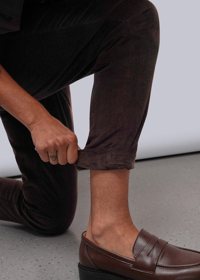 S=A close-up of a person wearing dark brown leather penny loafers with dark brown corduroy pants, kneeling on a gray concrete floor against a white studio background.
