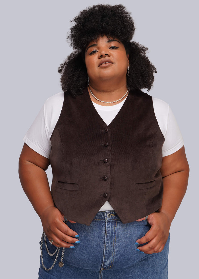 1X=A plus-size model wearing a dark brown corduroy vest over a white t-shirt and blue jeans, standing against a solid gray background.