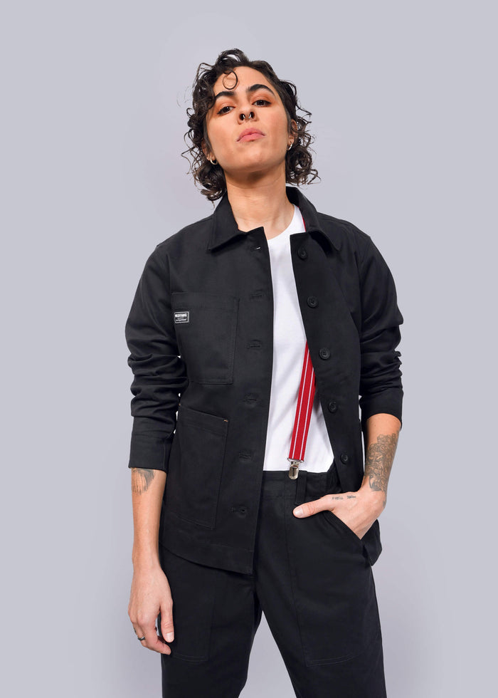 S=Wearing The Essential Utility Jacket and comfortable stretch pants, a person with short curly hair pairs their look with a white shirt and red suspenders, standing confidently against a white background.