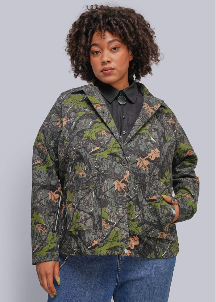 1X=A woman wearing a camouflage jacket with green, brown, and gray tones, styled over a black collared shirt and blue jeans, against a solid gray background.