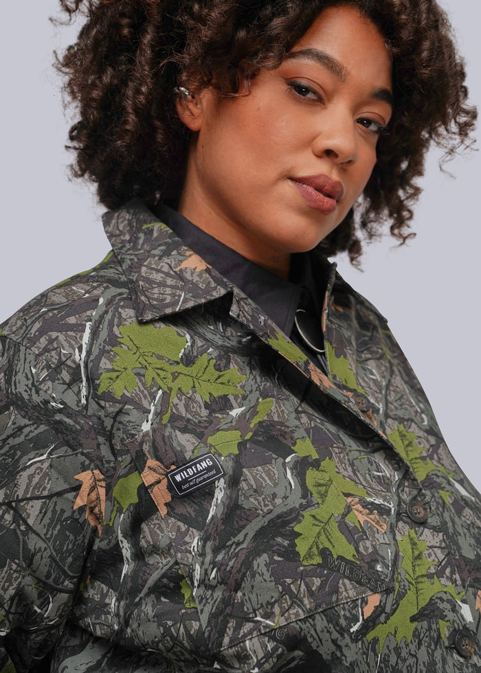 1X=A close-up of a model wearing a camouflage jacket with green leaf motifs and a black collared shirt underneath, featuring a visible 'WILDFANG' brand patch.