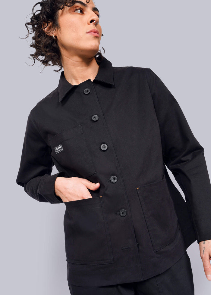 S=Wearing The Essential Utility Jacket in black, they stand against a plain background with their left hand in a deep front pocket. A small square label adorns the chest as their curly hair enhances the effortless blend of style and stretch comfort, while they gaze off to the side.