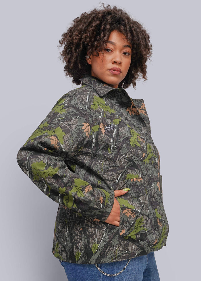 1X=A woman with curly dark hair models a camouflage jacket featuring green, brown, and gray woodland patterns, paired with blue jeans against a solid gray studio background.