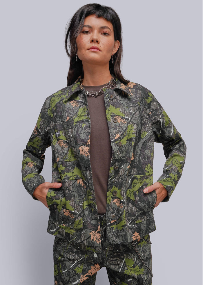 S=A model wearing a matching camouflage jacket and pants set over a brown top, posed against a solid gray studio background.