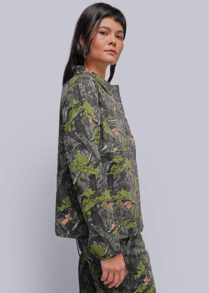 S=A model wearing a camouflage jacket and matching pants in a green, brown, and gray woodland camo pattern, photographed against a solid light gray background.