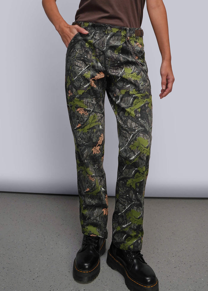 S=A model stands against a light gray studio background wearing a brown short-sleeve t-shirt, camouflage pants, and black boots.