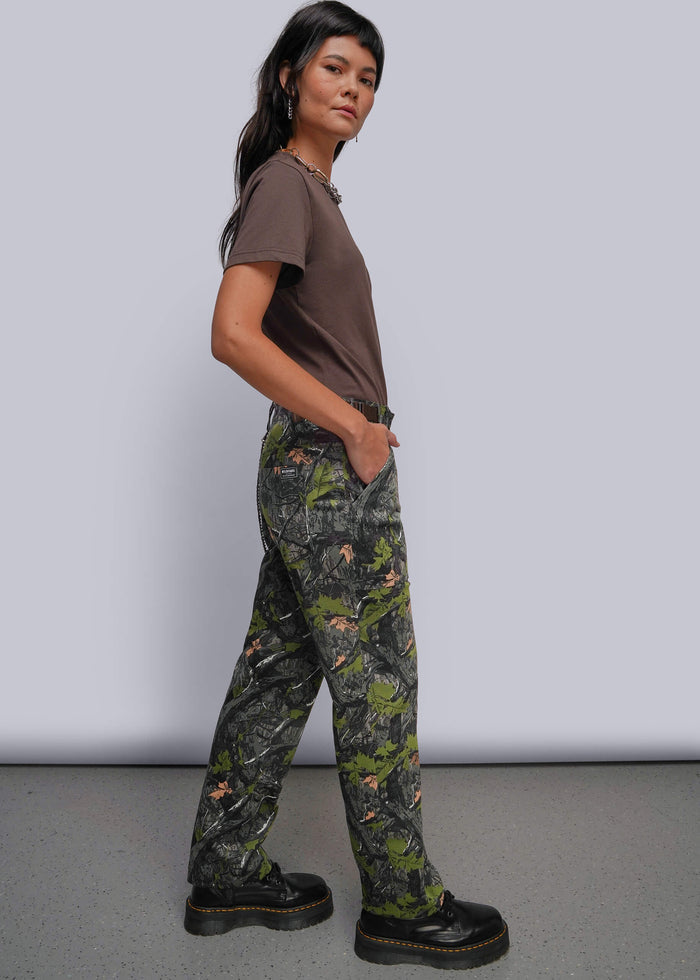 S=A model wearing a brown t-shirt and camouflage pants with a green and gray woodland pattern, paired with black platform boots, standing against a light gray studio background.