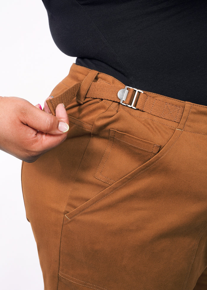 1X=Someone adjusts the waist strap on The Essential Utility Pant in brown stretch cotton, paired with a black top. The image highlights the hand and waistband detail, emphasizing comfort and fit.