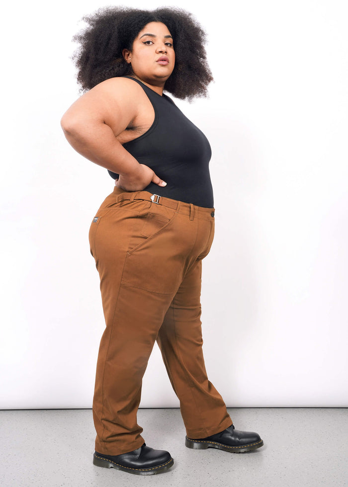 1X=A woman with curly hair models The Essential Utility Pant—rust-colored with roomy pockets and side buckles—paired with a sleeveless black top and black shoes, standing sideways against a white background.