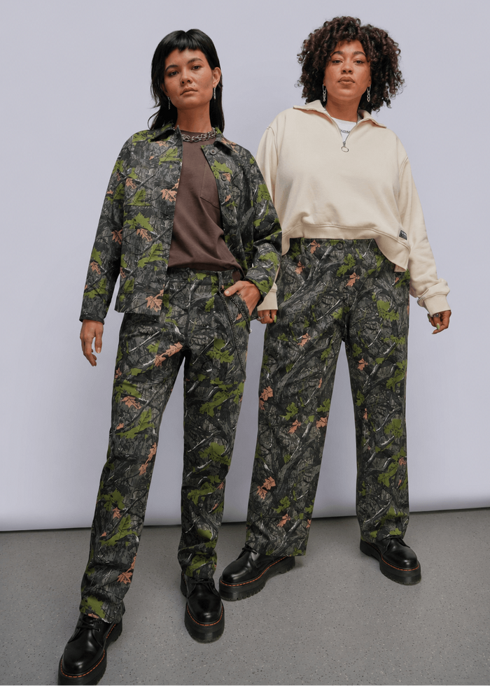 Two models standing side-by-side in a studio setting. The model on the left wears a matching camouflage print jacket and trousers set with a brown shirt underneath and black boots.