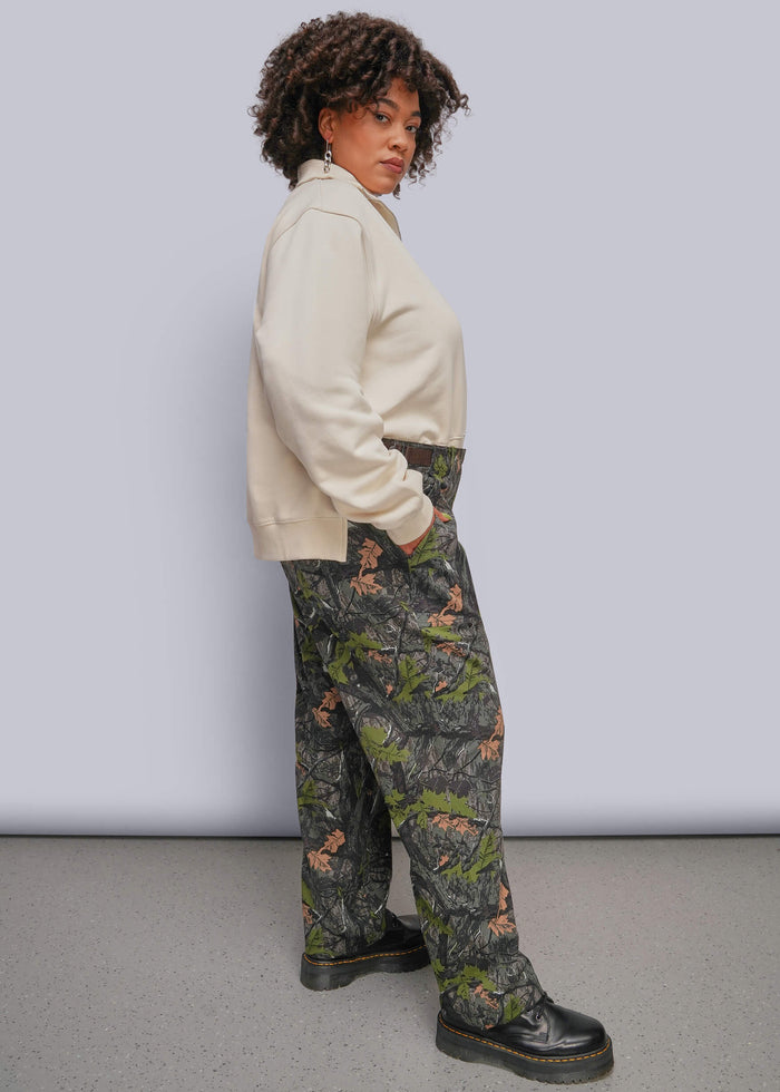1X=A model wearing a cream sweatshirt and camouflage pants with a green and brown woodland print, paired with black boots, standing against a light gray studio background.