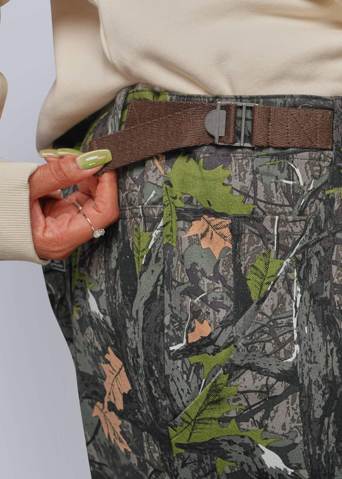 1X=Close-up of a person wearing camouflage pants showing adjustable waist with brown belt and metal buckle, a cream sweatshirt, and a hand adjusting the belt.