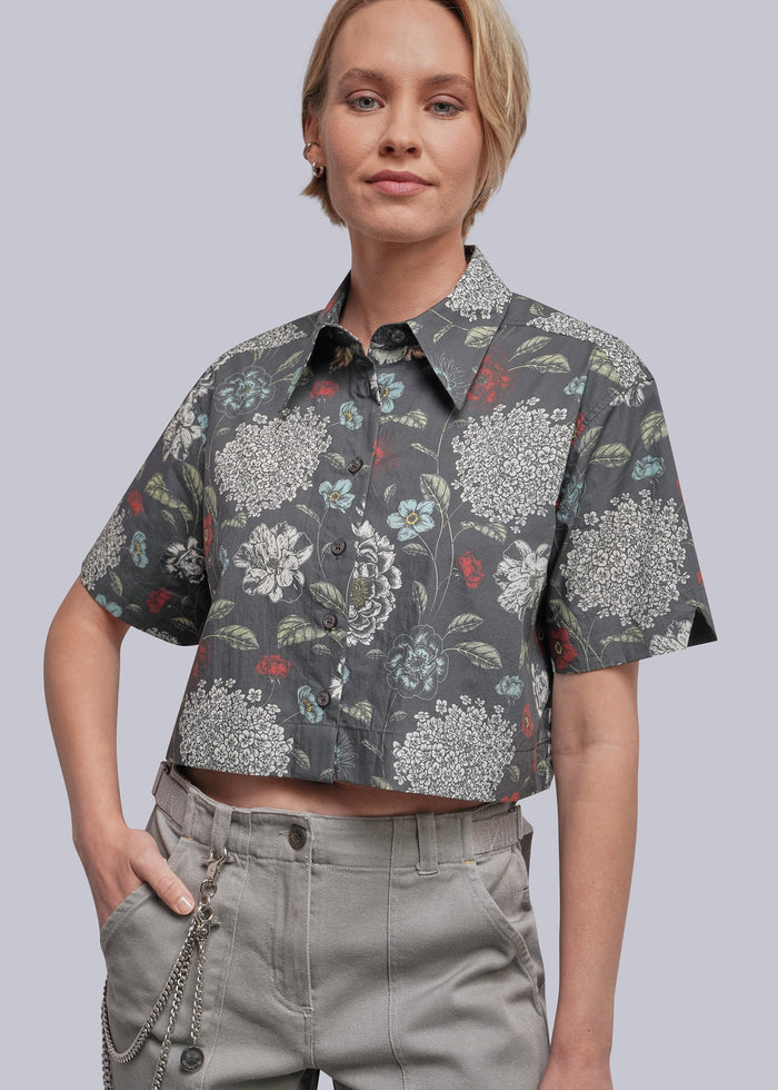 S=A woman wearing a cropped short-sleeve button-up shirt with a dark gray floral print and light gray pants, photographed against a plain light gray studio background.