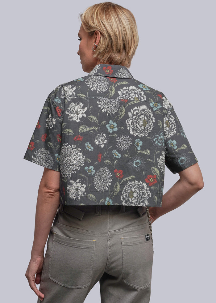 S=A woman shown from the back wearing a short-sleeve button-up shirt with a dark gray base and a white, green, and red floral pattern, paired with olive green pants, against a solid gray background.