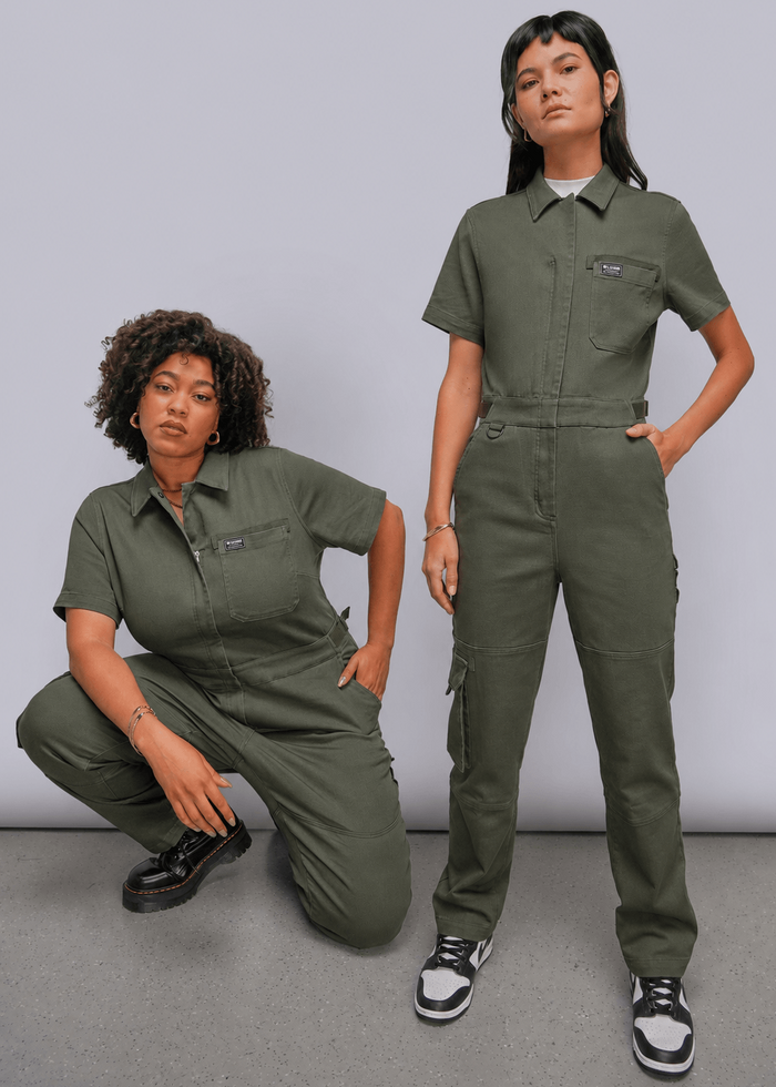 Two models wearing matching olive green short-sleeve utility jumpsuits, posed against a neutral light gray studio background.