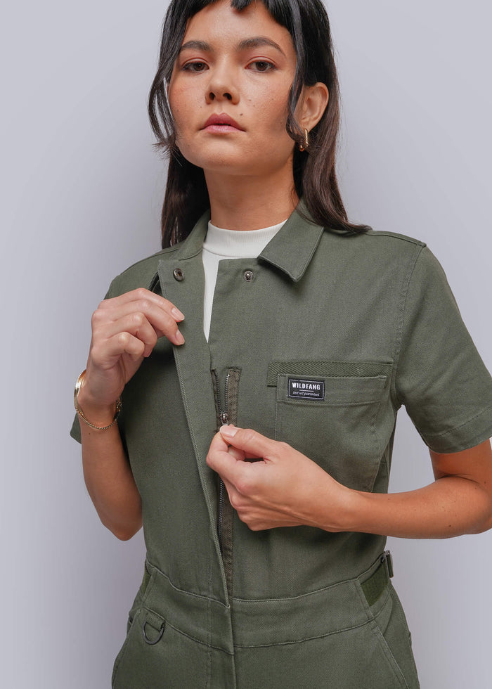 S=A woman wearing an olive green short-sleeve utility jumpsuit with a chest pocket and a black 'MILLIEE' brand label, styled over a white t-shirt against a light gray studio background.